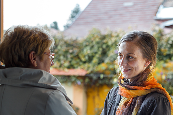 A young woman in a yellow scarf greets an older woman, at the front of a house