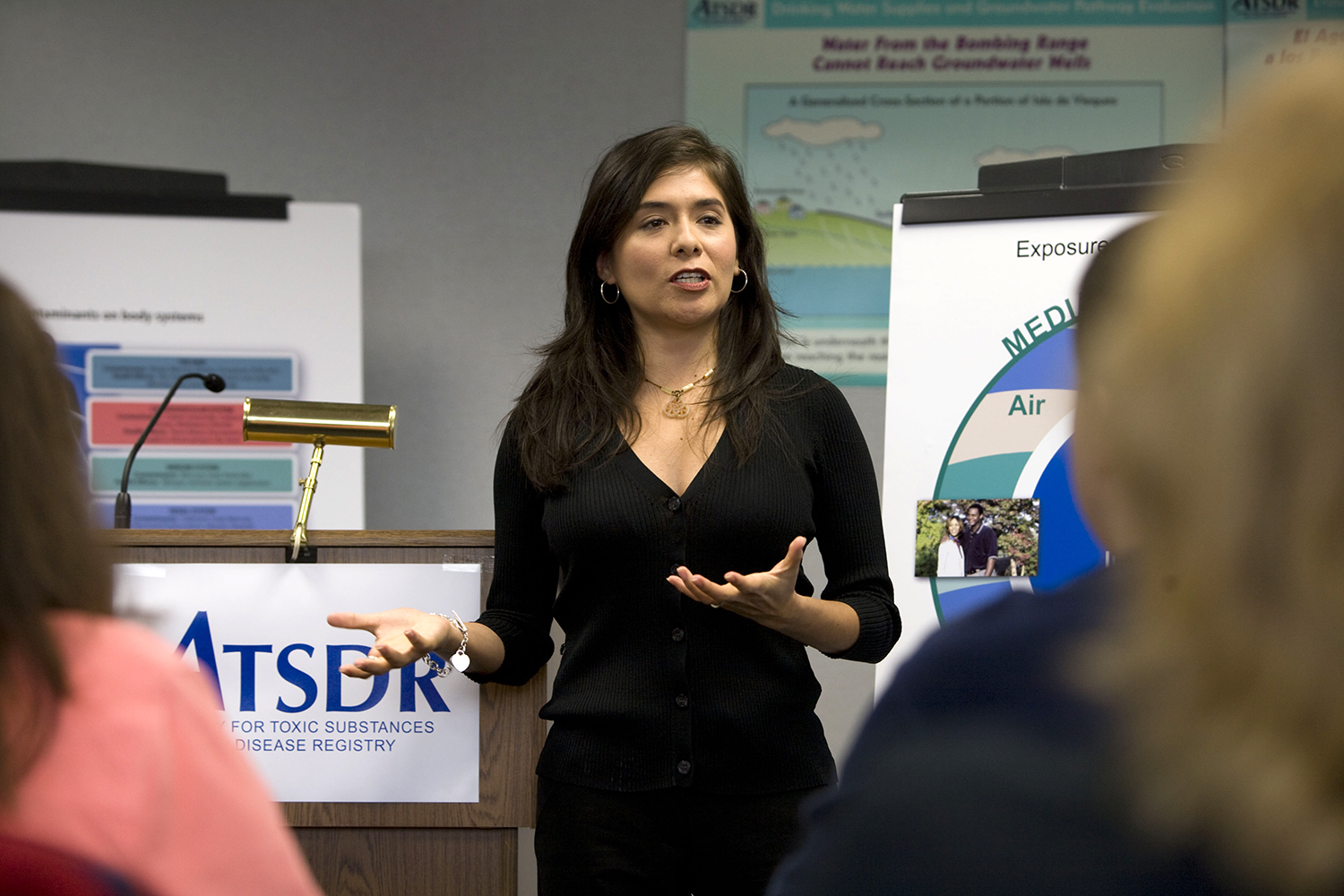A Hispanic woman speaks to attendees at a town hall meeting in front of a podium with an ATSDR sign displayed.