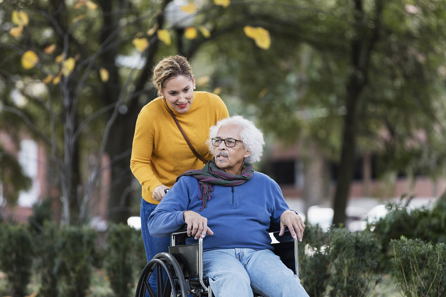 A senior Hispanic man in his 80s sitting in a wheelchair, enjoying a walk in the park with his adult daughter, an adult woman in her 30s.