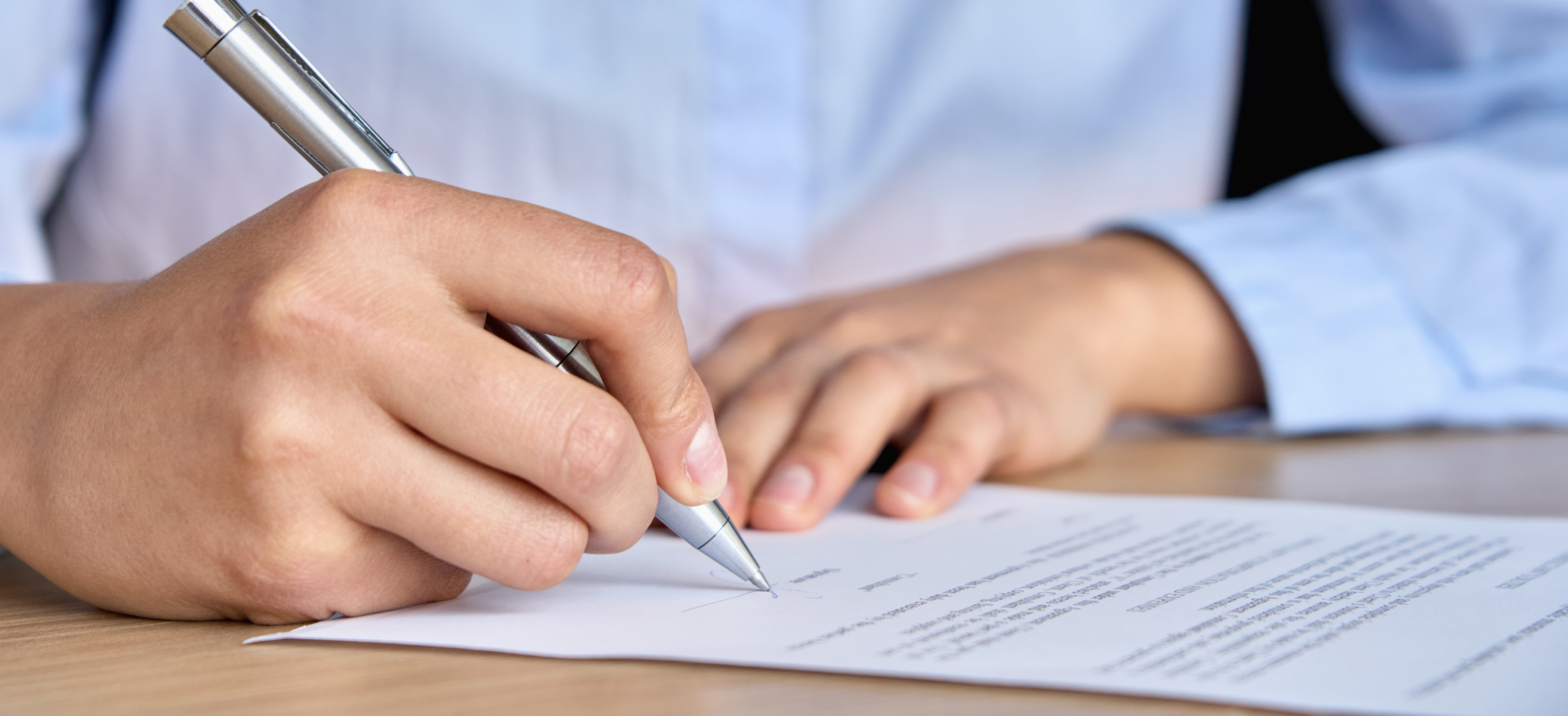 A person wearing a blue shirt is signing a piece of paper using a silver pen.
