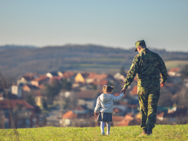 A man wearing military attire is walking and holding a little’s girl’s hand in a field with houses in the background.