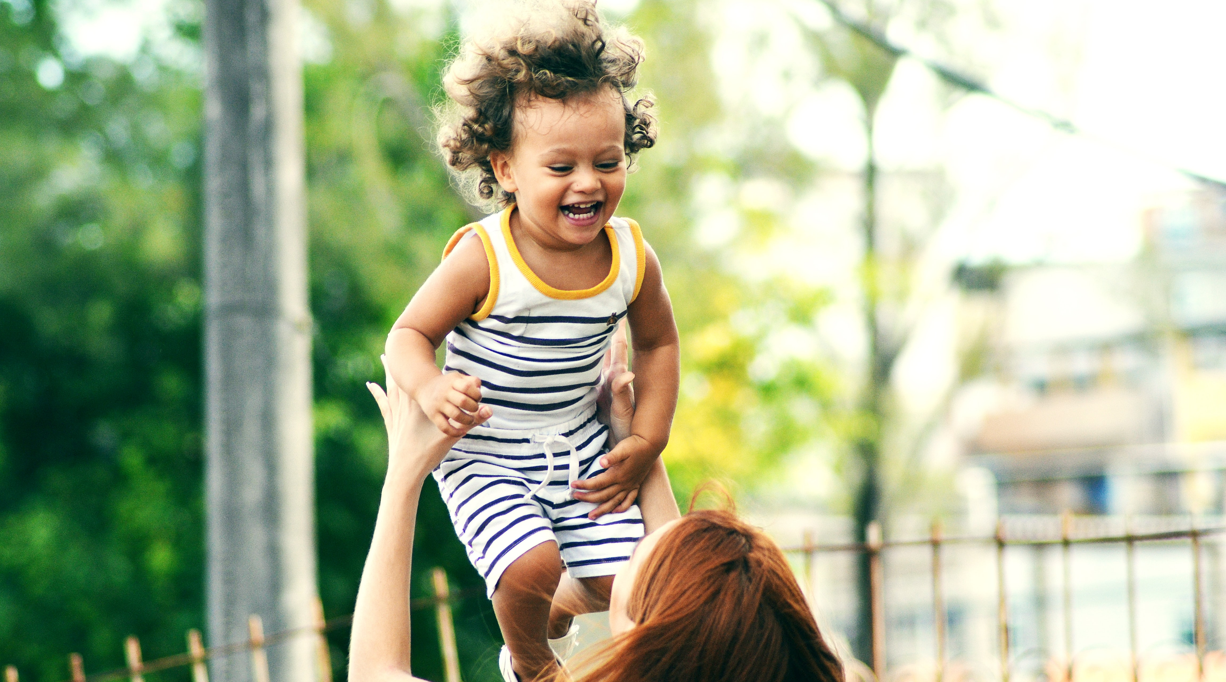 A laughing child with a striped outfit and curly hair is being tossed in the air by a woman.