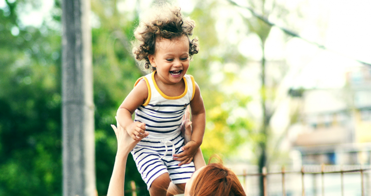 A laughing child with a striped outfit and curly hair is being tossed in the air by a woman.