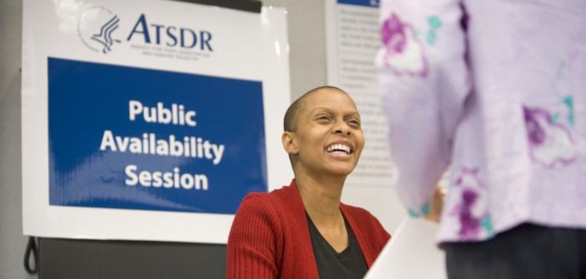 A smiling woman is looking up at a person during a meeting. Behind the smiling woman is a sign that says ATSDR public availability session.
