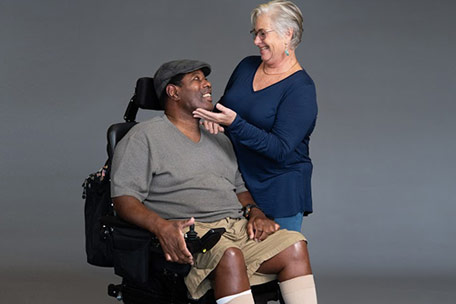 A man with ALS in a wheelchair exchanges a smile with a caregiver.