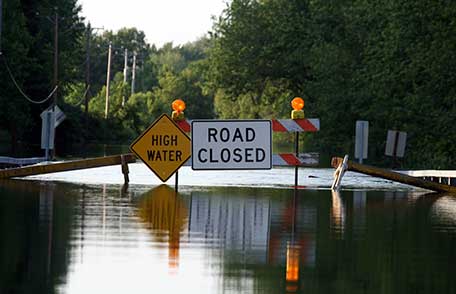 Una calle residencial inundada con señales de High Water y Road Closed que bloquean la entrada.
