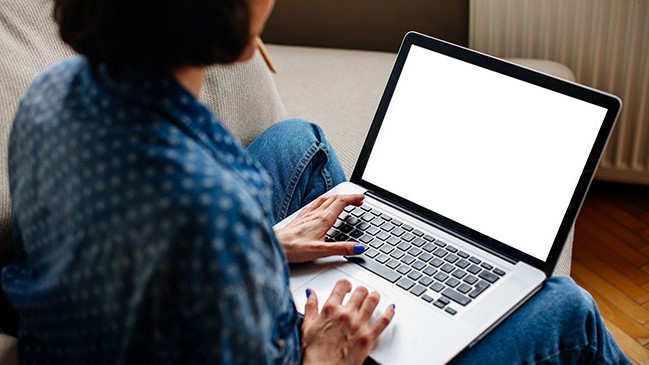 PFAS Blood Estimation Tool A woman with a blue and white shirt and blue jeans sitting on a couch looking at a blank laptop screen.