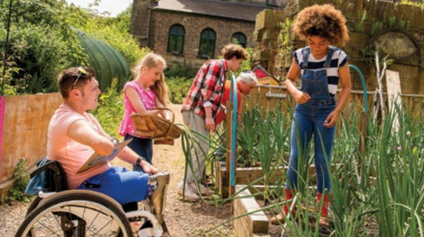 Group Gardening A man in a wheelchair, a girl in overalls and boots, and others outside in a garden.