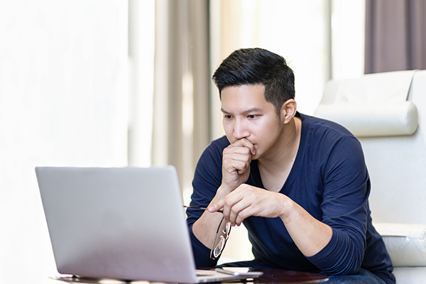 Young man working with his glasses in one hand and leaning his head in his other hand while looking at a laptop.