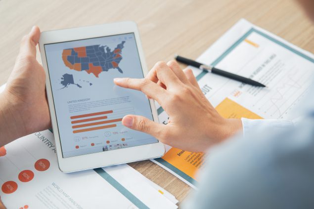 Person at a desk holding a tablet that has a map of the United States and a bar graph.