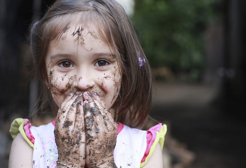 A smiling child. The child’s hands are covering her mouth and there is dirt and mud on her face.