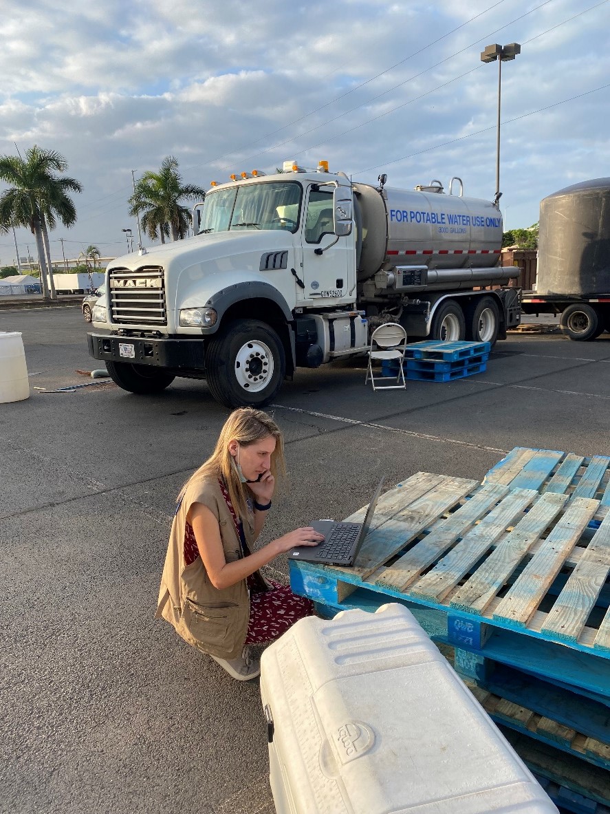 ACE investigation team member works on her laptop on a makeshift desk of stacked pallets next to a potable water truck.