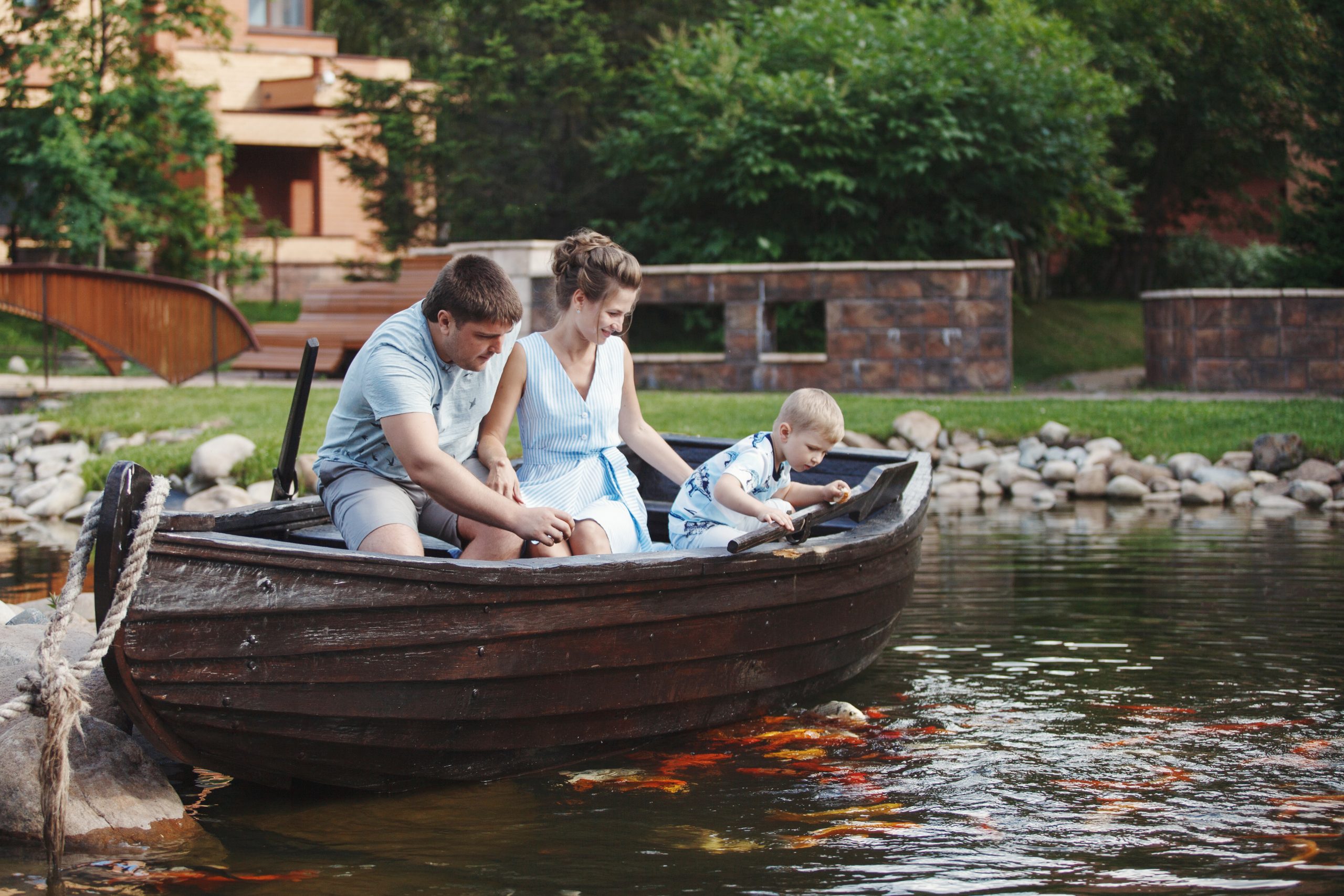 Mother, father and son sit in a wooden boat with oars and feed the fish with bread.